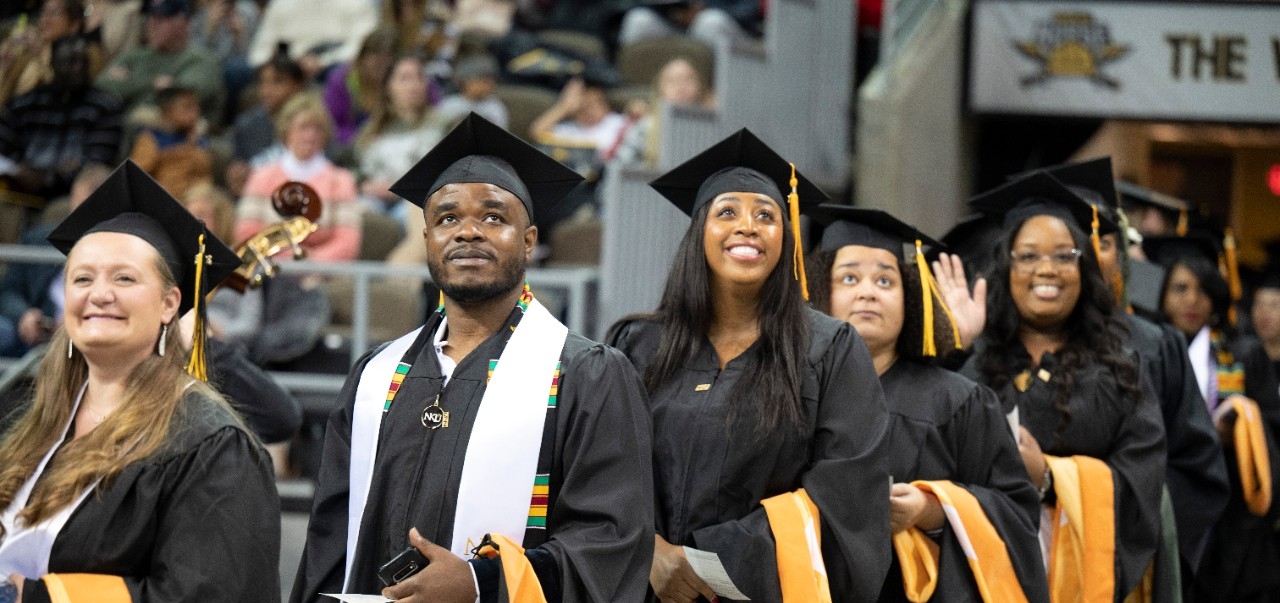 A line of students enter Truist Standium for the Fall 2022 commencement ceremony. Three black students look up at the families in the stands. 