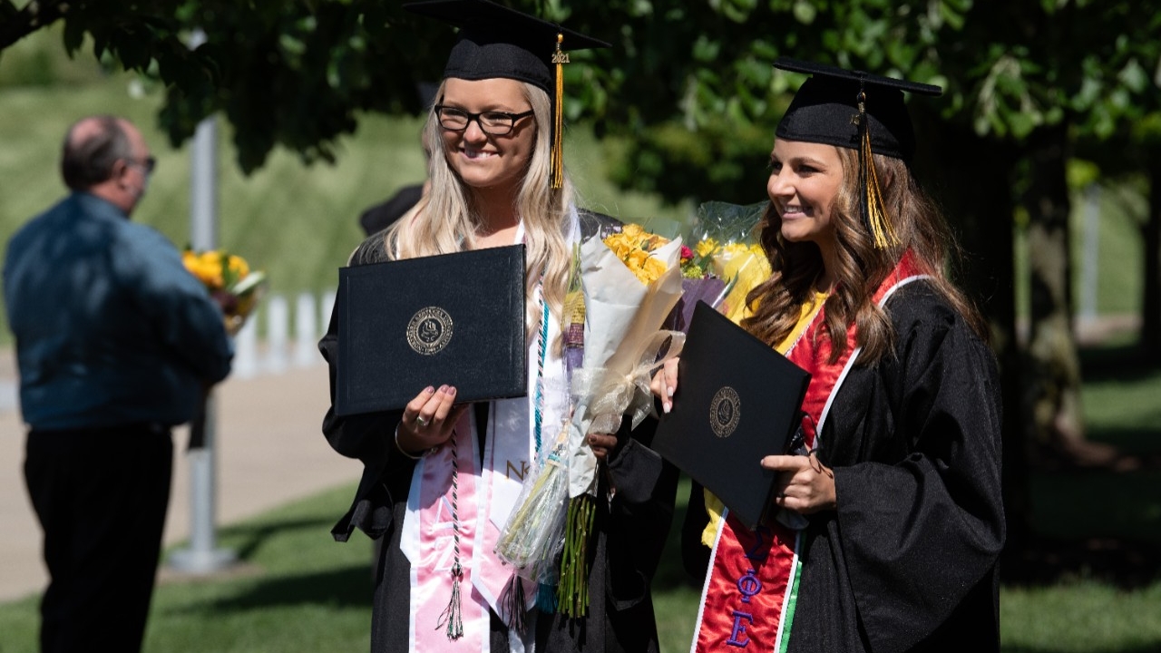 Two female graduates holding their diplomas outdoors.