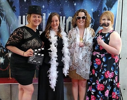 Four people standing behind a Hollywood background holding props.
