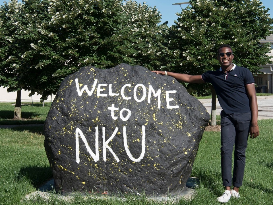 Student standing next to a large rock that has the words "Welcome to NKU" painted on