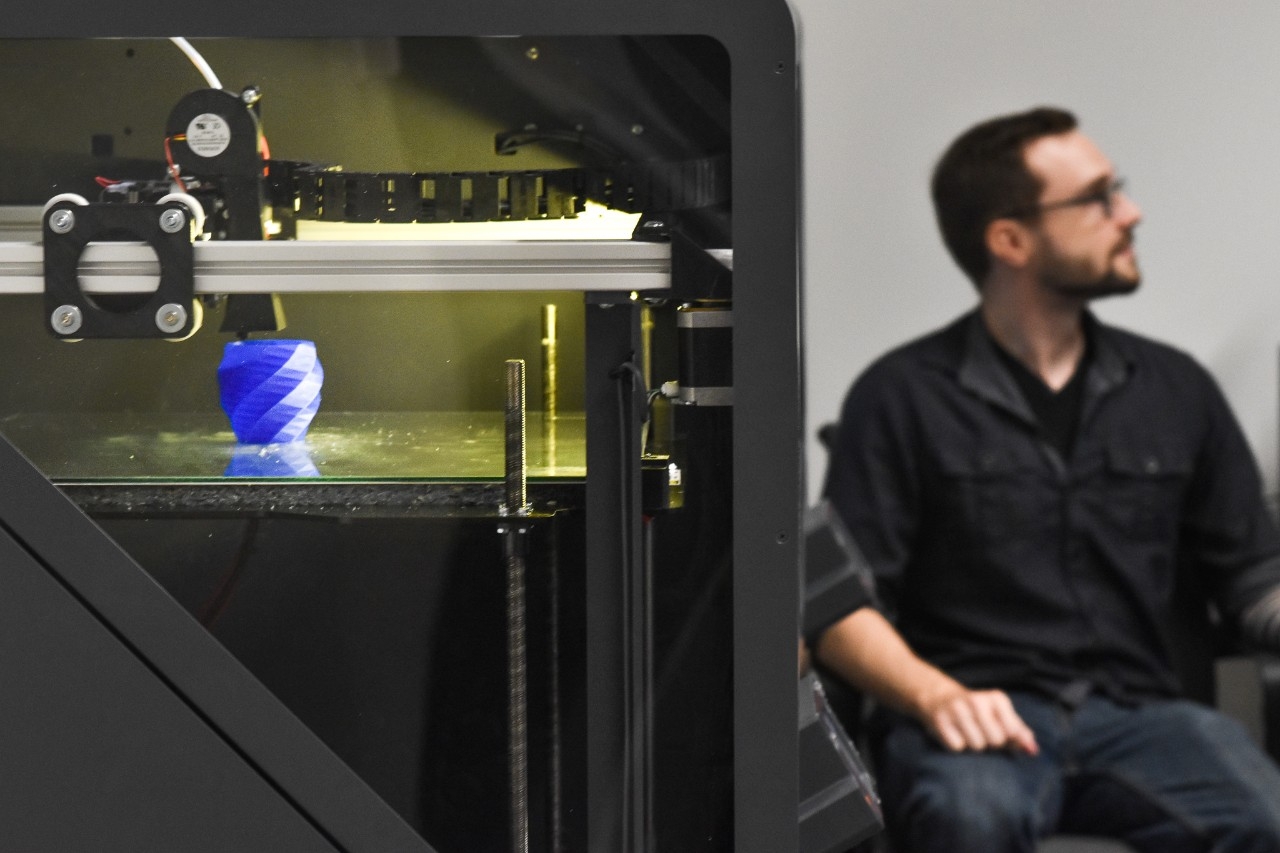 Vase being 3D printed in a large 3D printer with a man sitting in the background.