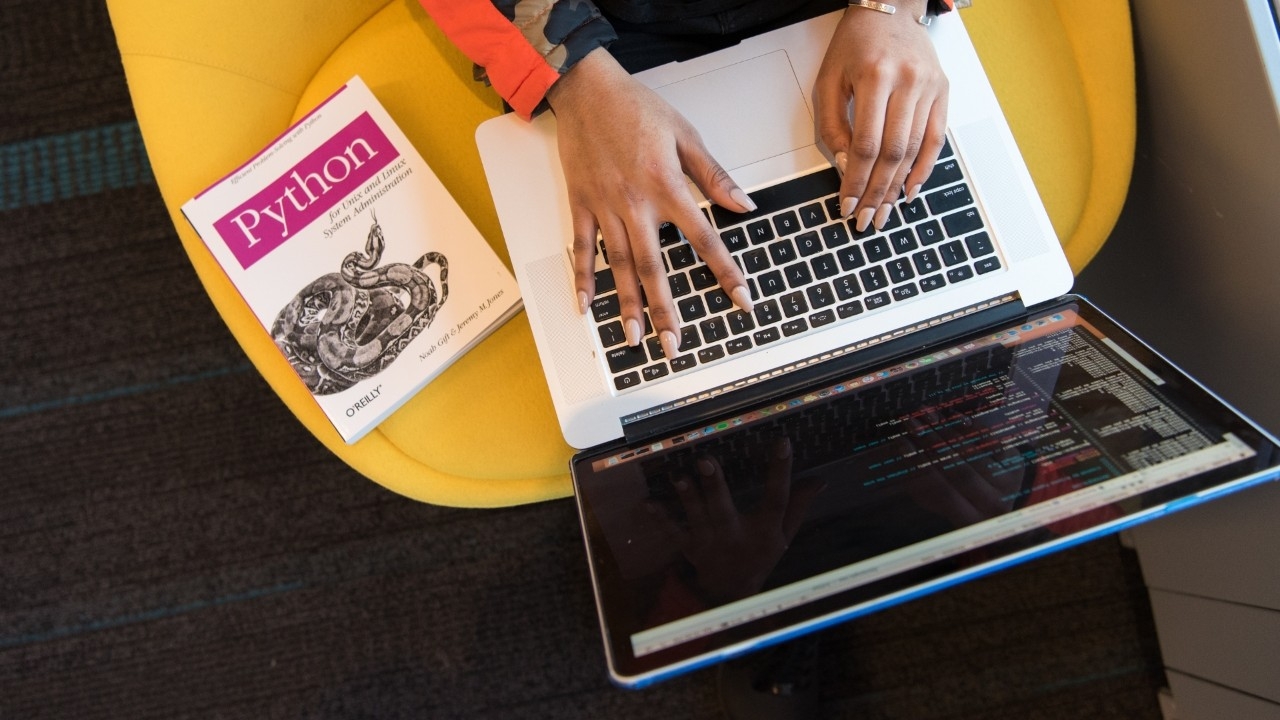 Woman sitting on yellow chair while working on laptop with a book next to her. 