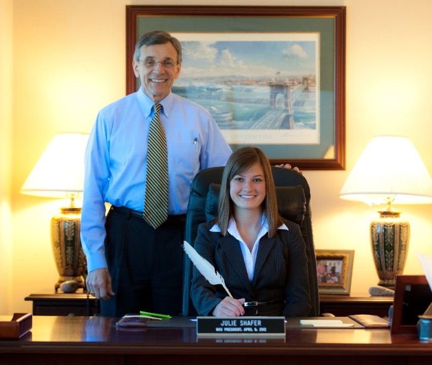 Two people smiling; one sitting at desk and one standing behind the desk