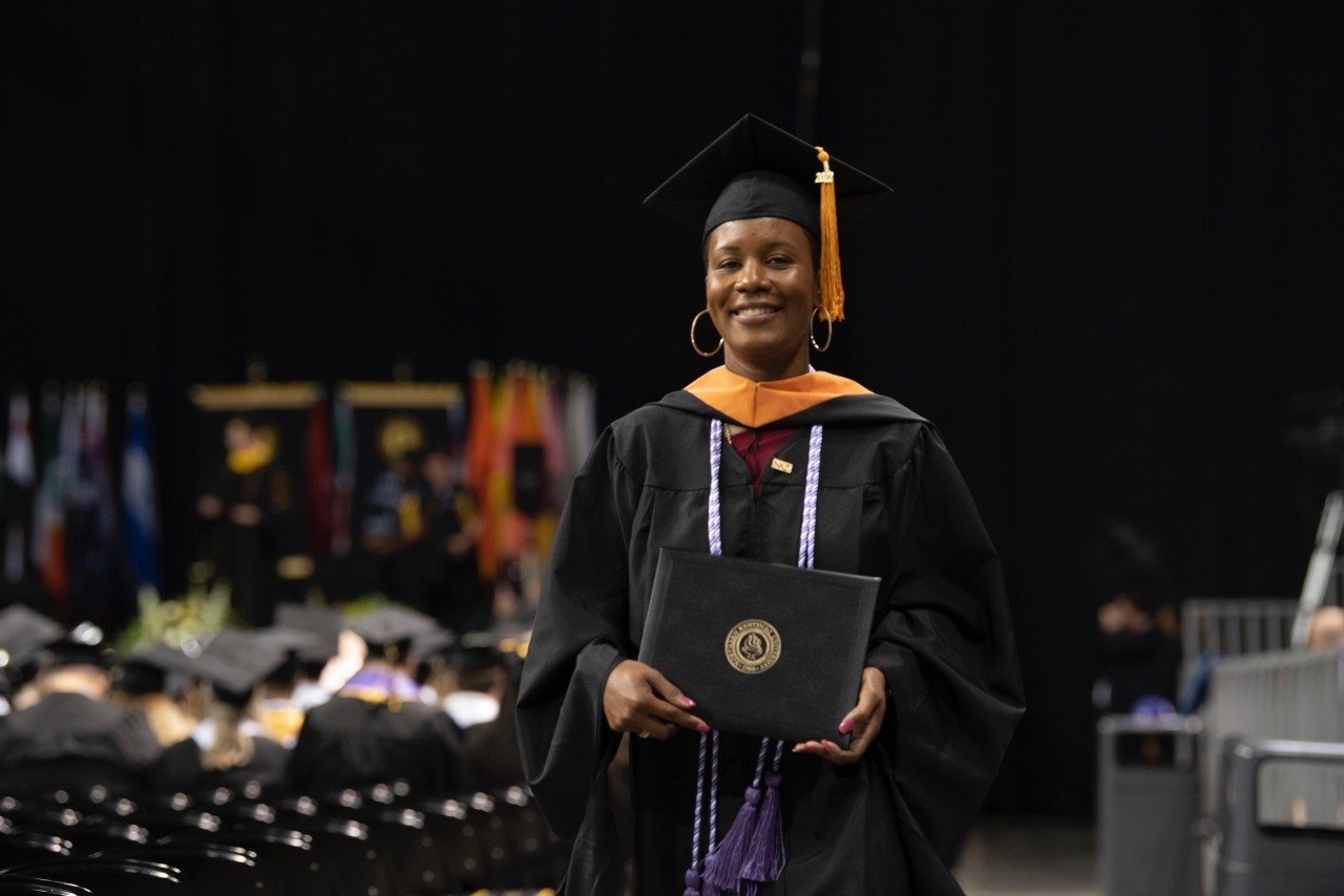 A female black student at Commencement smiles as she holds her diploma. 