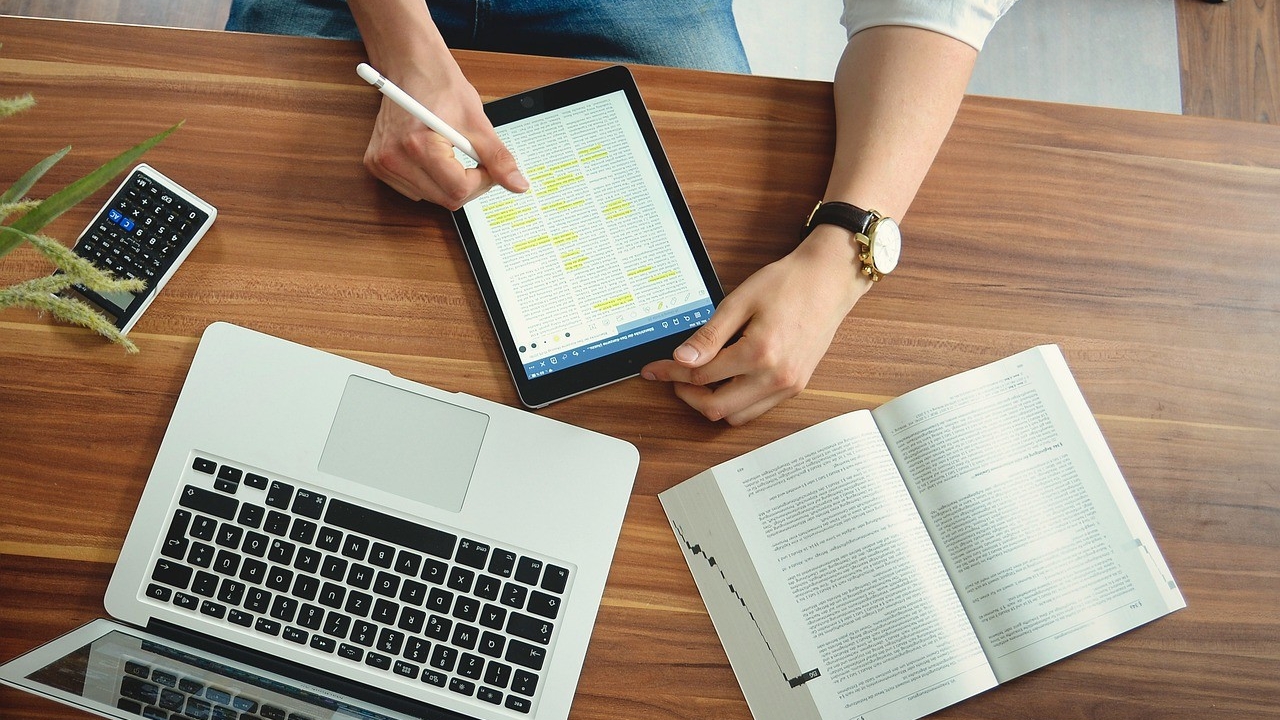 Someone working on a tablet in front of an open book and computer laptop