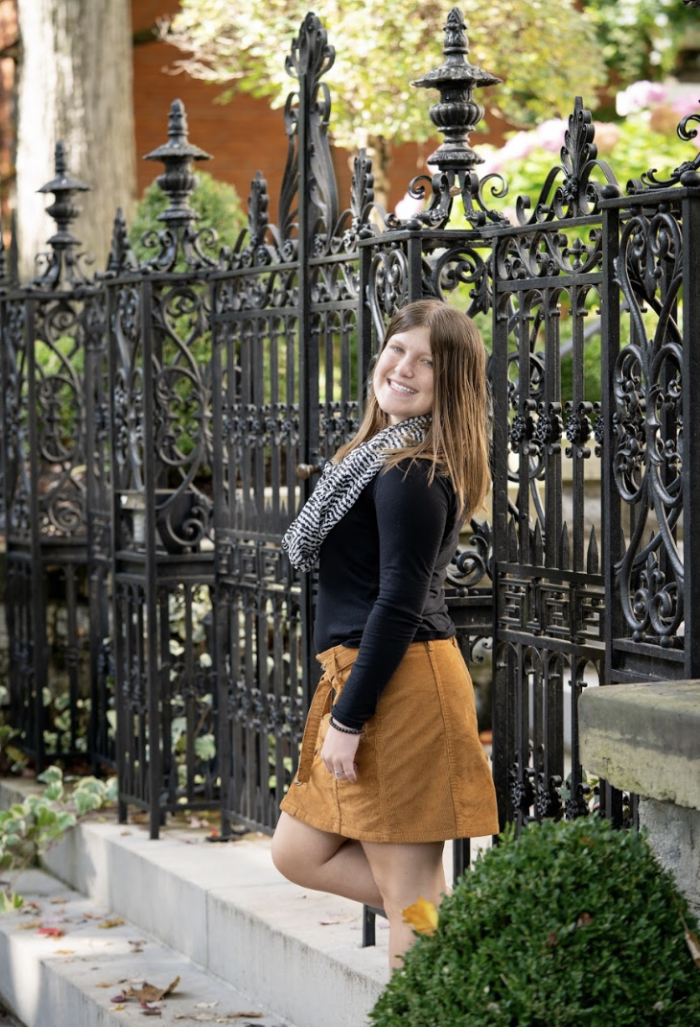 Photo of Julia in a black shirt and tan skirt with greenery and an iron fence in the background