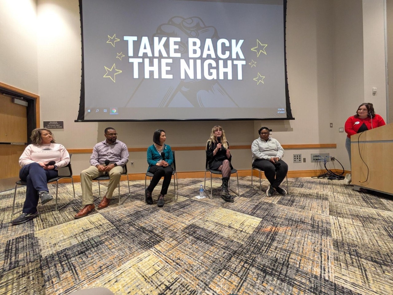 Five individuals sitting on a panel under a screen with Take Back the Night logo.