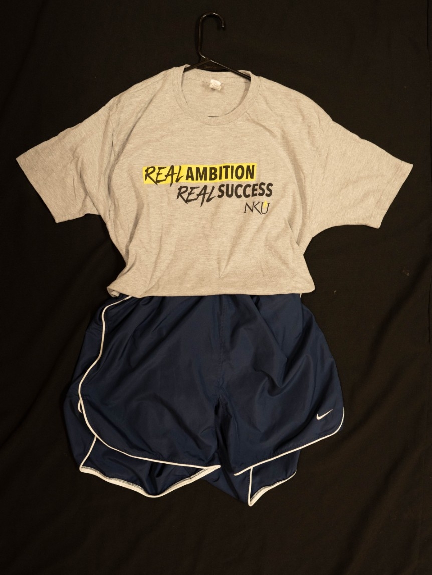 Exhibit displays an NKU t-shirt with “Real Ambition, Real Success” across the center with blue athletic shorts. 