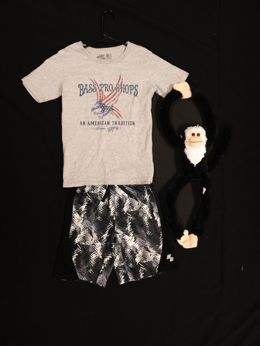 Exhibit displays a gray child’s t-shirt, black camo child's shorts, and a stuffed animal monkey. 