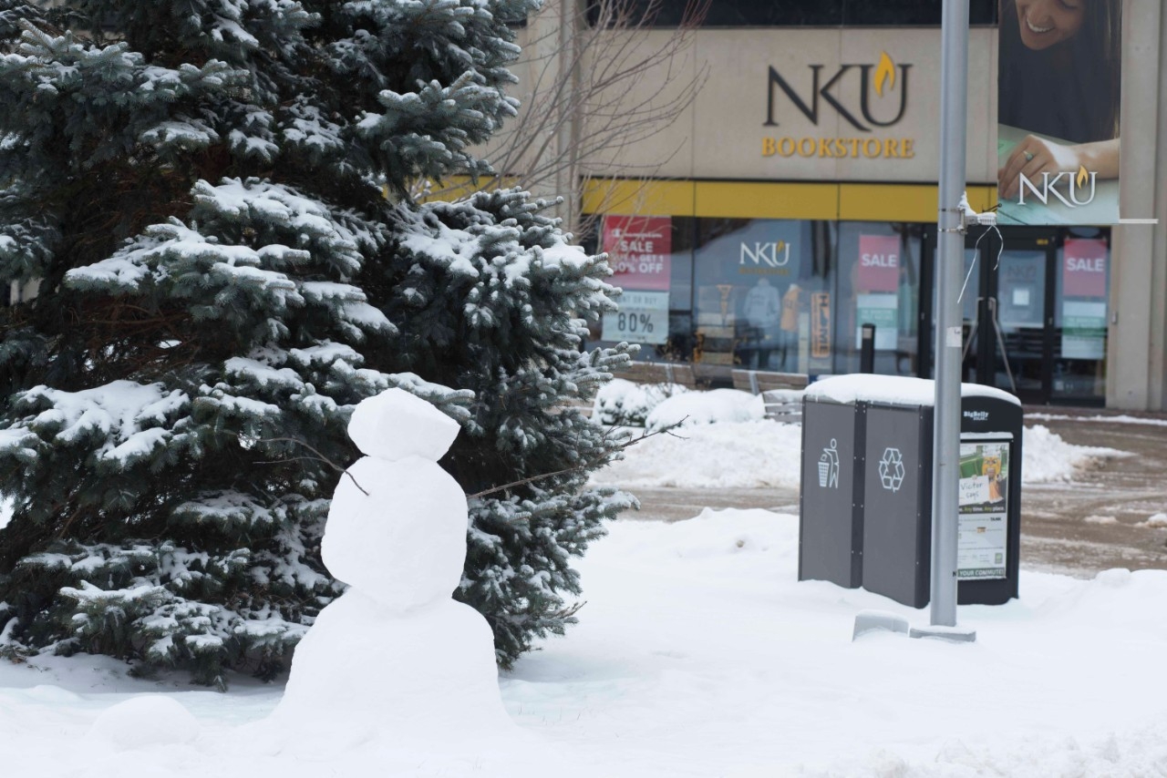 A snowman built in front of a snowy tree and the NKU Bookstore