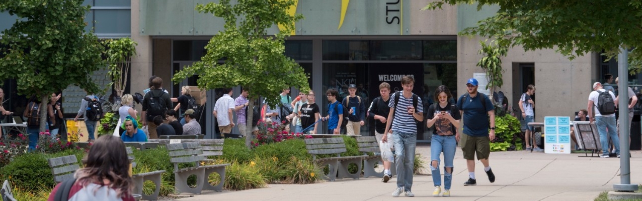 Many NKU Students walking outside of the Student Union, pictured with full trees on a warm day