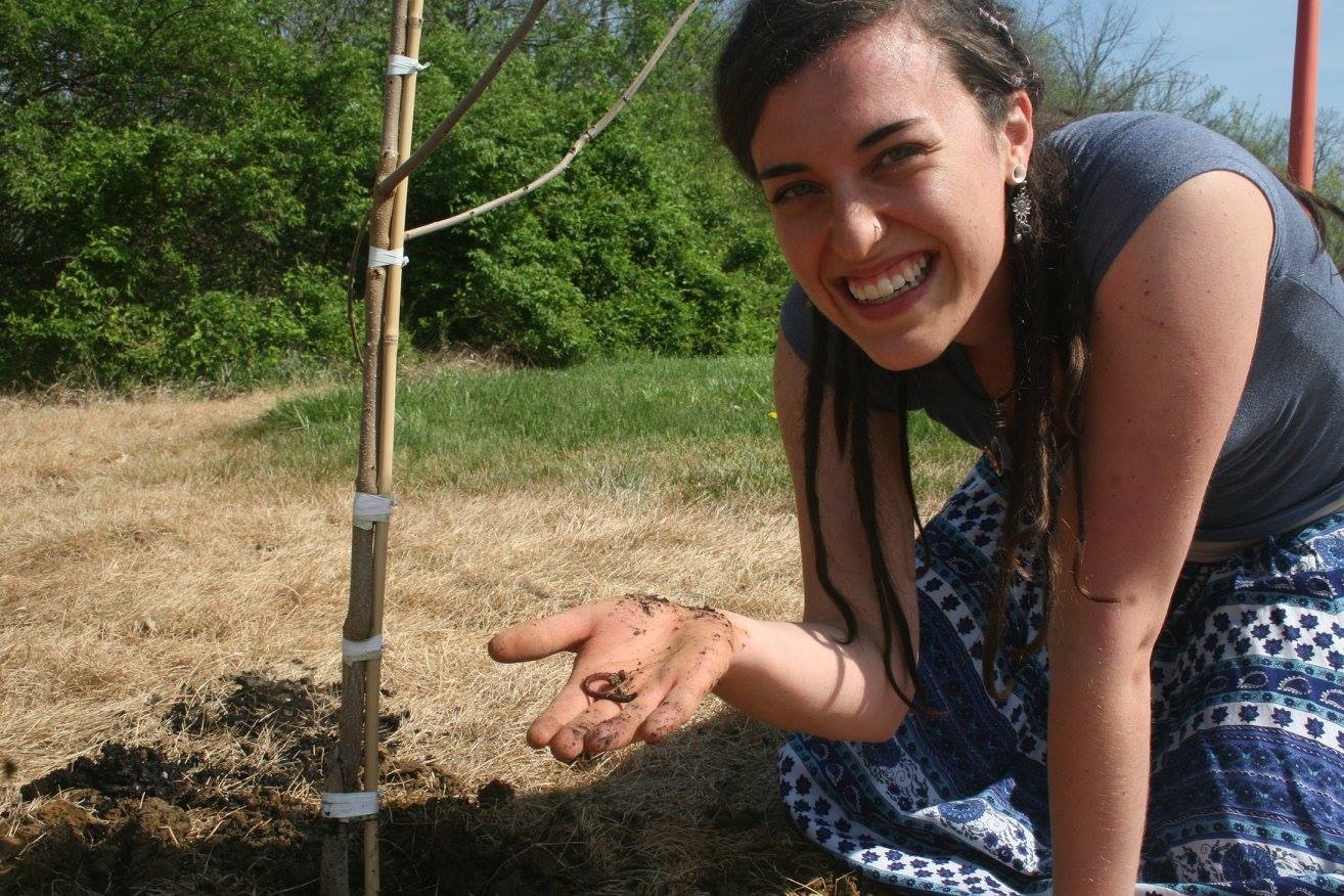 A gardener with long hair and printed skirt kneels on the ground holding a worm