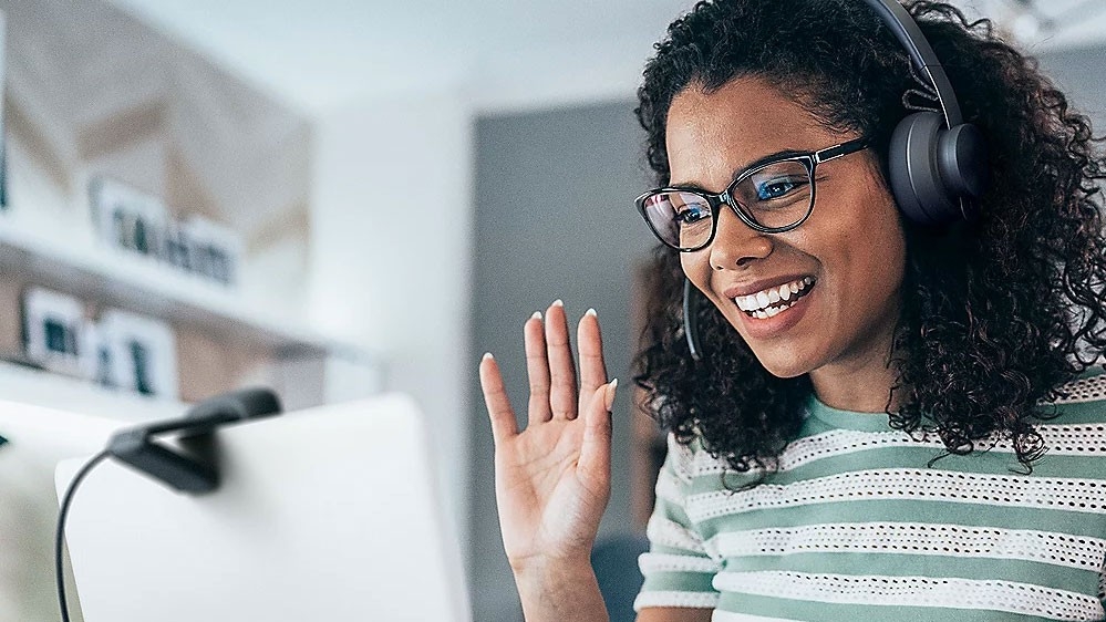 Woman wearing headphones, smiling and waving at her camera.