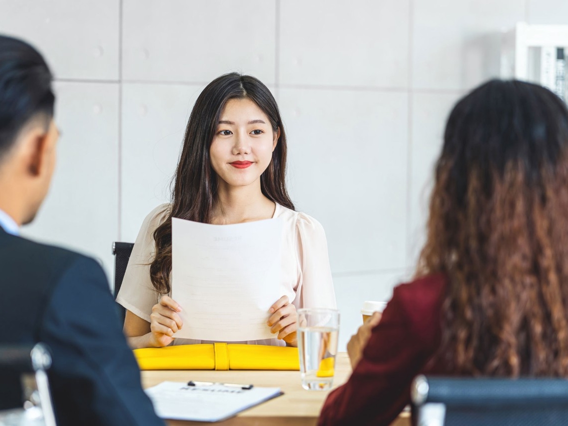 A young woman holds up her resume while facing two interviewers.