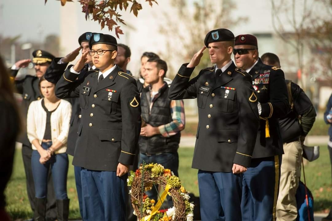 ROTC students gathered together, saluting at wreath ceremony taking place in a cemetery. 