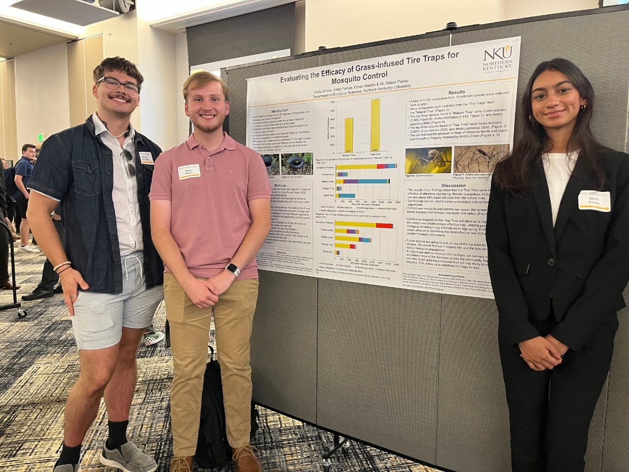 Three NKU students smiling in front of a poster presentation of their research